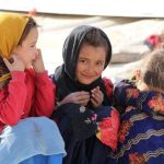 A group of young girls are sitting on the ground in front of tents.